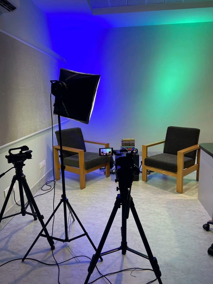 A small interview set with two chairs, a softbox and two cameras on tripods under blue and green light.